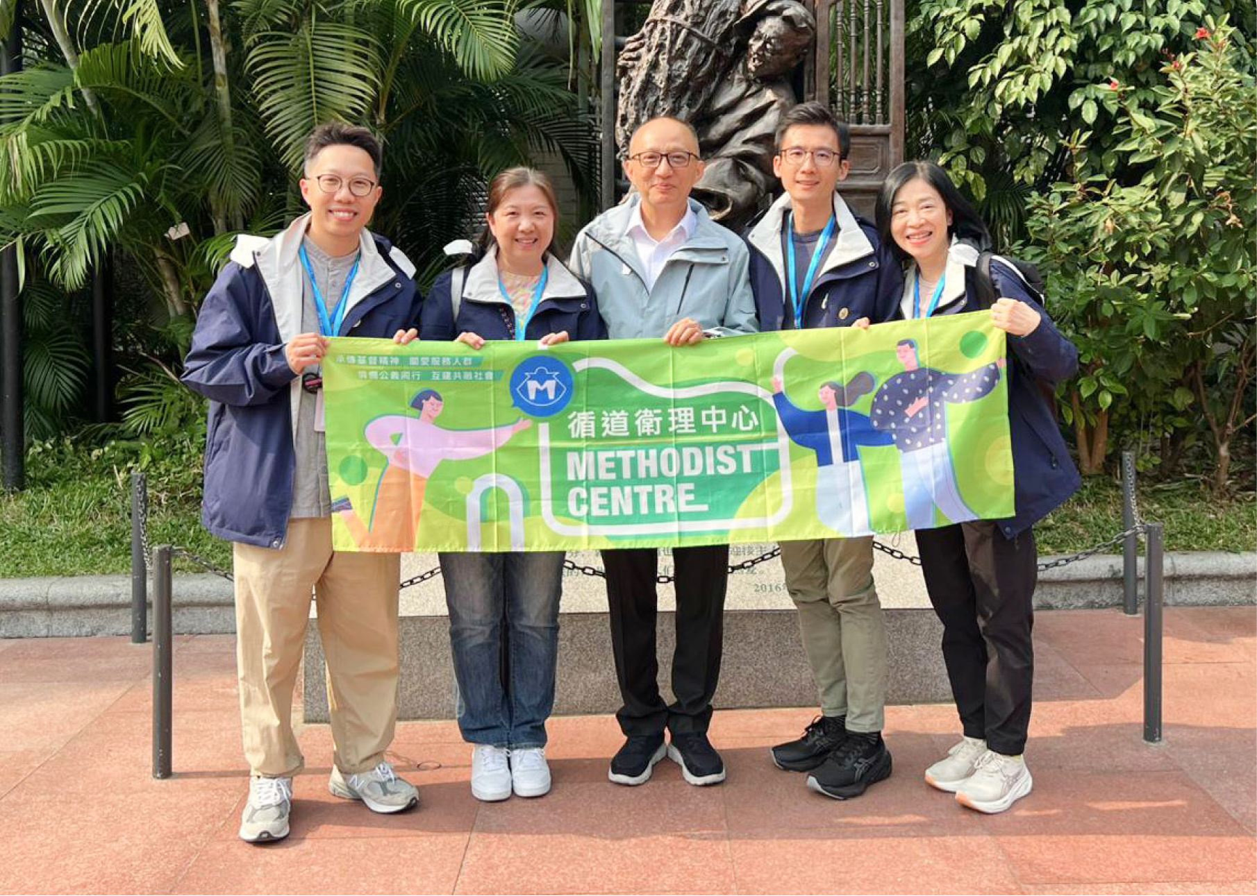 循道衛理中心職員於戶外雕塑前舉起中心橫幅合影